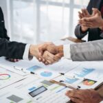 Business men shaking hands with numbers on papers under their hands.